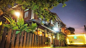 A traditional wooden house with a balcony, illuminated by warm lights, surrounded by lush greenery.