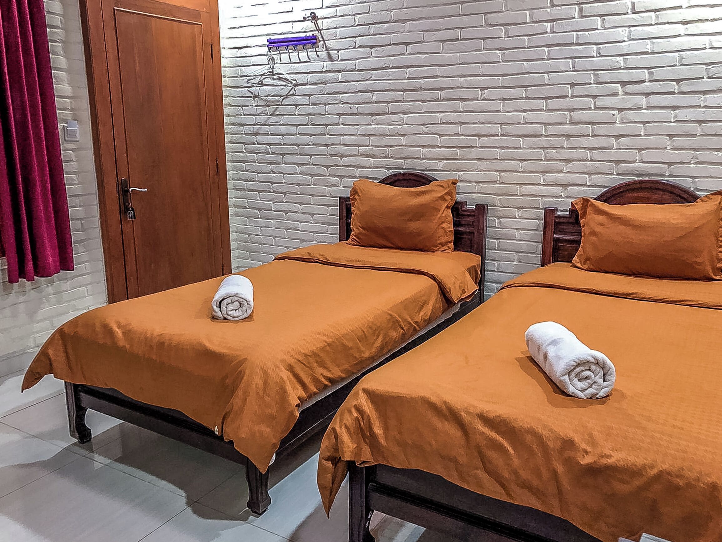 Une chambre avec deux lits simples, chacun recouvert d’une couverture marron et agrémenté d’oreillers, une armoire en bois et un mur en briques avec une tringle pour suspendre des vêtements.