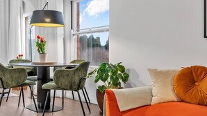 Una sala moderna con una mesa de comedor redonda, sillas tapizadas de verde, una lámpara colgante negra, una planta grande en maceta y un sofá naranja con un cojín.