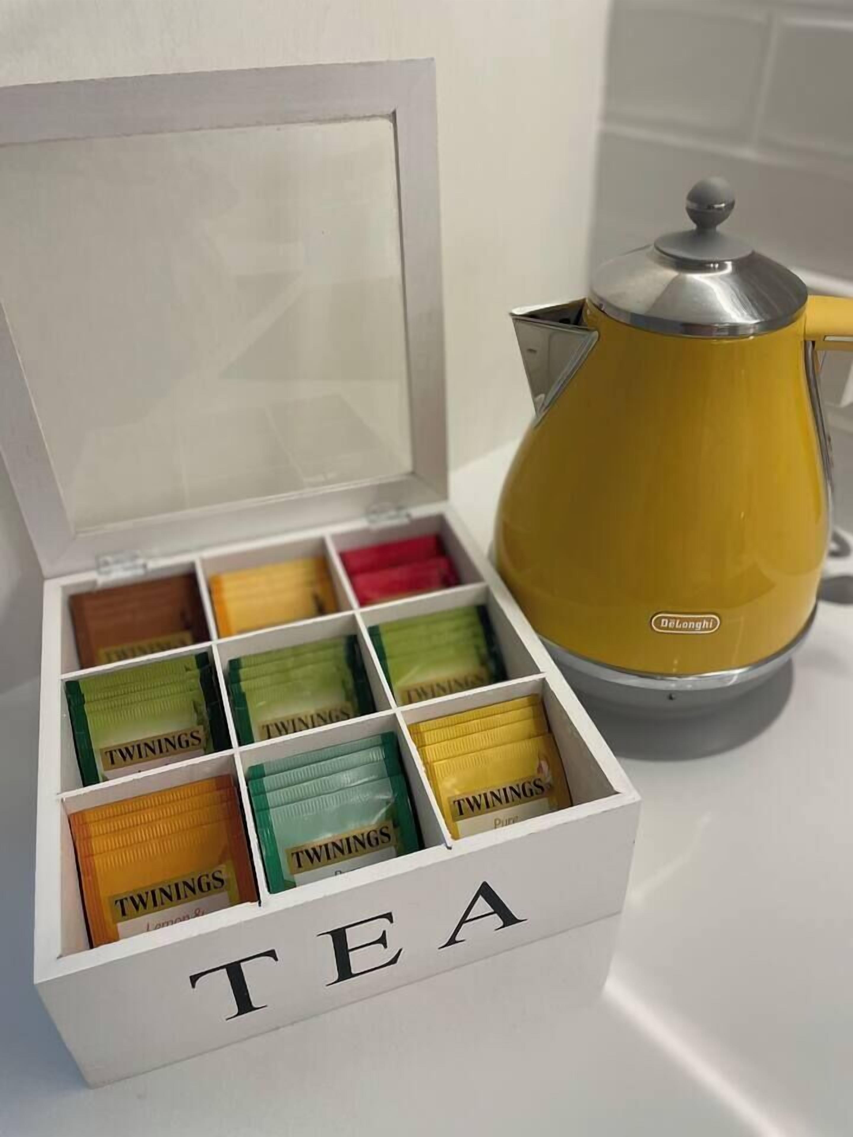 A white box labeled 'TEA' with compartments containing tea packets, next to a yellow DeLonghi coffee maker.