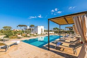 A swimming pool with lounge chairs, a covered seating area, and a white building in the background.