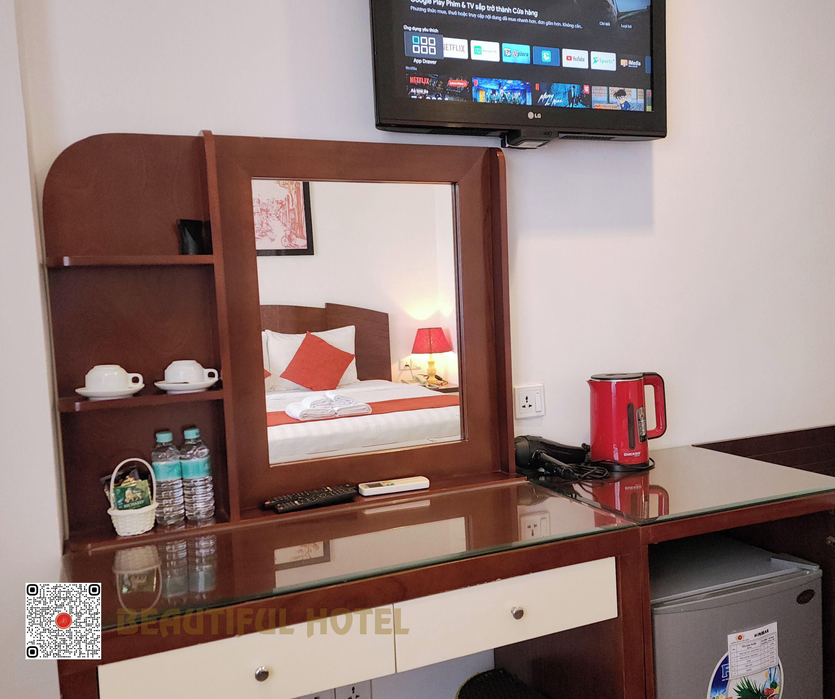 Habitación de hotel con escritorio de madera, cama con almohada roja, televisor montado en la pared y tetera roja sobre el escritorio.
