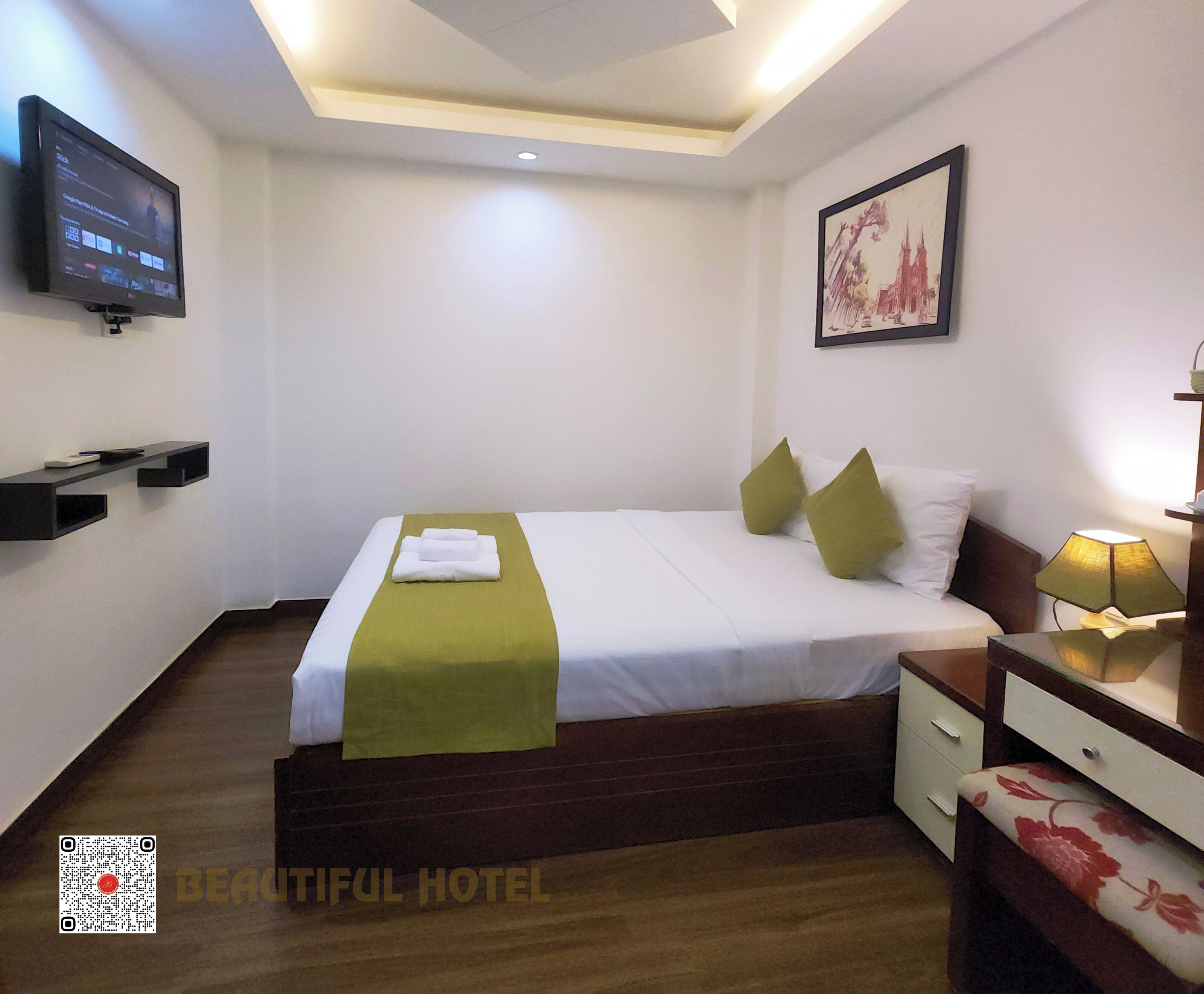 Una habitación de hotel con una cama, un televisor montado en la pared, una mesita de noche con una lámpara y un cuadro enmarcado.