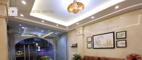 A hotel lobby with a brown leather sofa, a round wooden coffee table, potted plants, framed artwork on the wall, and a fan.
