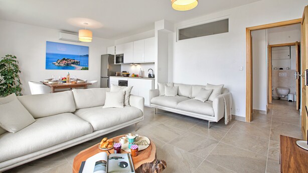 Una sala de estar moderna con un sofá, una mesa de centro y un comedor con mesa y sillas. Se observa una cocina al fondo con armarios y una encimera.