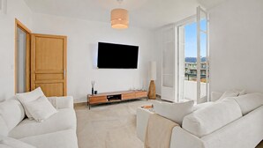 Una sala moderna con una televisión de pantalla plana montada en la pared, una puerta de madera, un ventanal corredizo que da a un balcón y un sofá color beige.