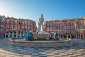 A public square with a central fountain, featuring statues and a building with multiple stories and balconies.