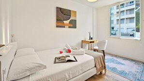 Habitación de hotel con cama, escritorio, silla, ventana con vista a edificios y un cuadro en la pared.