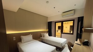 A hotel room with two beds, a desk, a chair, an air conditioning unit, and a view of the hallway. - Shankee Sudirman (Jakarta)
