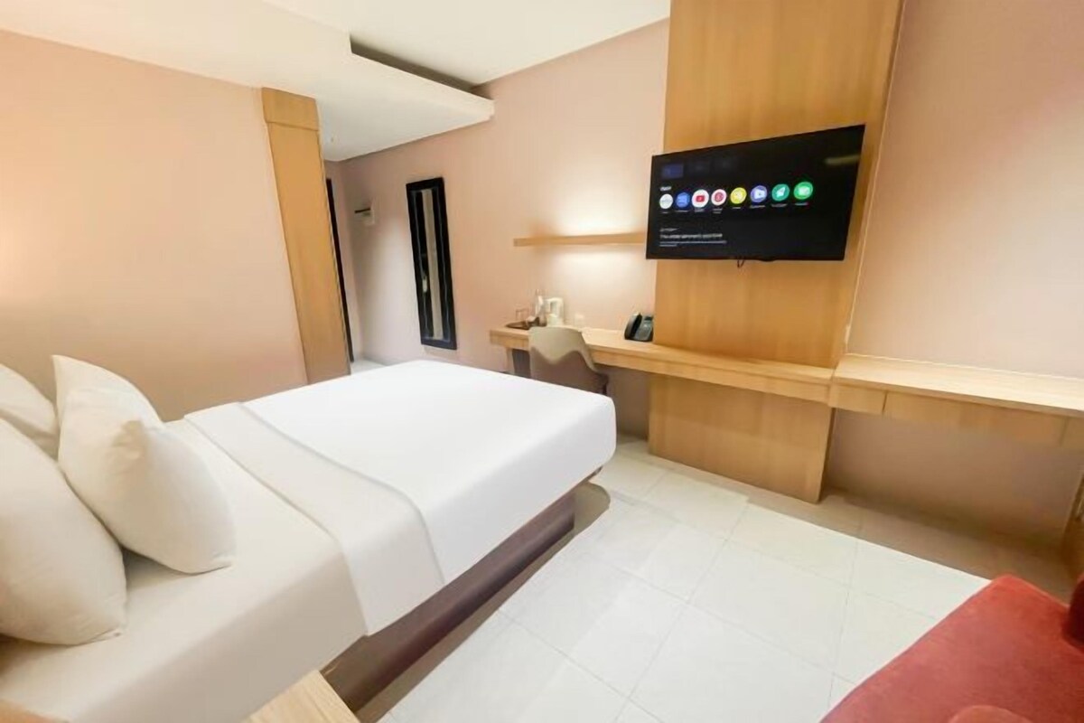 A modern hotel room with a bed, a desk, a flat-screen TV mounted on the wall, and a small seating area with a red chair.