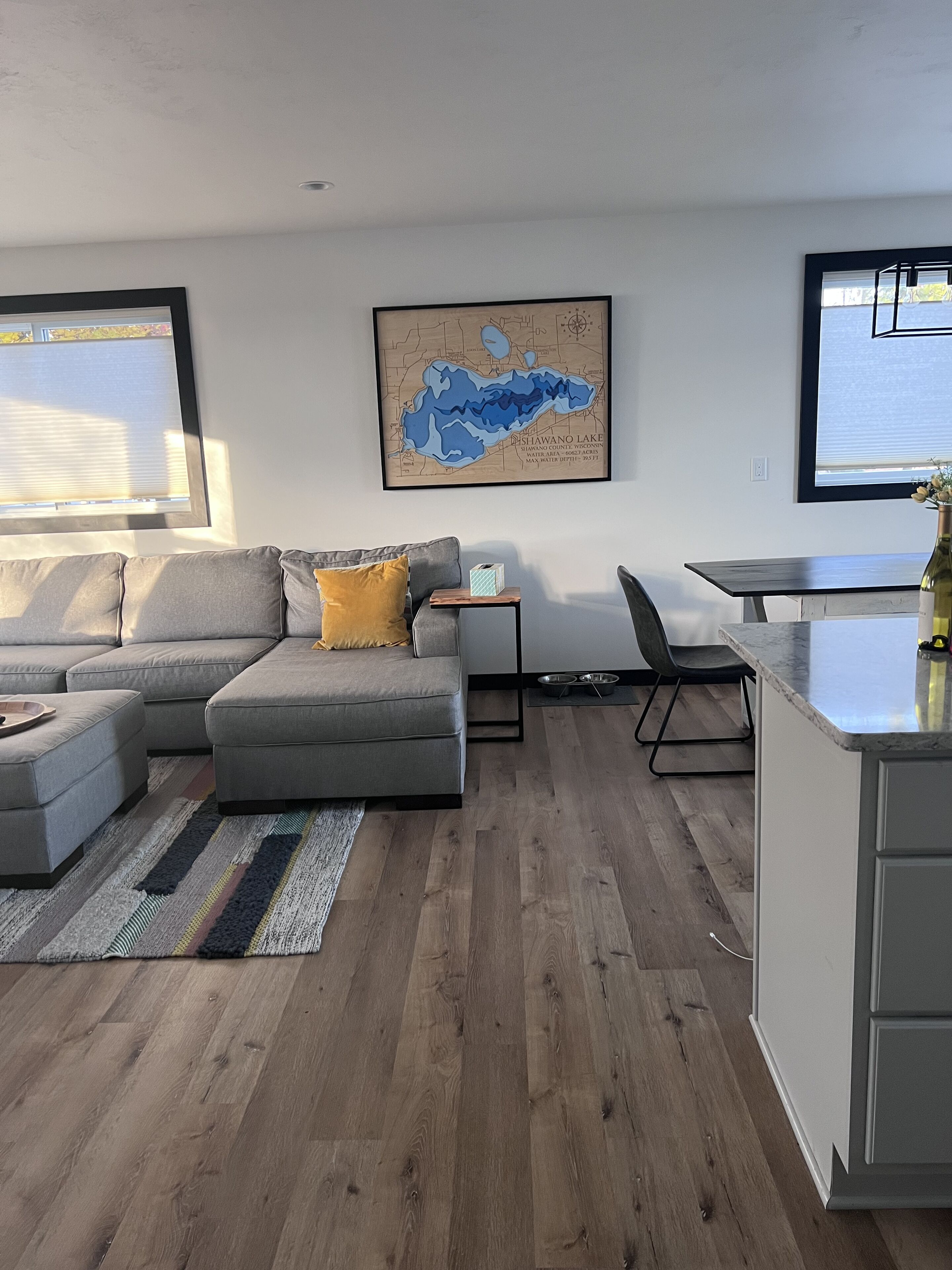 Una sala moderna con un sofá seccional gris, una pequeña mesita de madera y un mapa del lago Shawano en la pared.