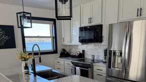 A modern kitchen with white cabinets, a stainless steel refrigerator, a built-in microwave, and a granite countertop. There are three wooden stools at the island. - Over 200’ of  lakefront all sand on Washington lake with a view of Shawano lake! (Shawano)