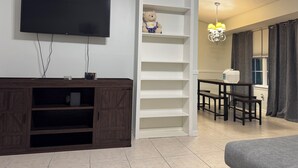 A living room with a TV, a wooden entertainment unit, a staircase, a dining area, and a chandelier.