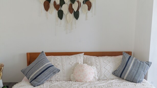 Una cama bien hecha con edredón blanco y almohadas estampadas, cabecera de madera y un adorno colgante en la pared.