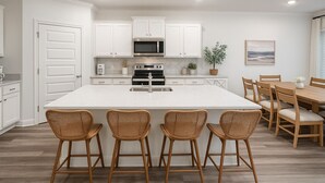 A modern kitchen with white cabinetry, a central island with bar stools, a dining table with chairs, and a view of the ocean through the window. - Sleeps 10 - The Salty Piper - Community pool (Santa Rosa Beach)