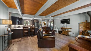 A spacious living area with a kitchen, wooden flooring, and a large TV mounted on the wall. - Escape to Tranquility in This Beautiful Forest-Edge Retreat (Graeagle)