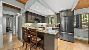 A modern kitchen with a central island, dark cabinetry, stainless steel appliances, and a view of the outdoors. - Escape to Tranquility in This Beautiful Forest-Edge Retreat (Graeagle)