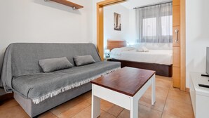 Una habitación moderna con un sofá gris, una mesa de centro de madera, una cama con cabecera de madera y un televisor sobre un mueble blanco.