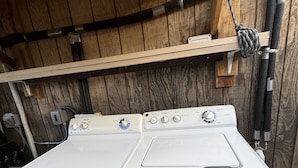 Lavadora y secadora frontales blancas en un cuarto de lavado de madera con tuberías visibles y un estante metálico en la parte superior.
