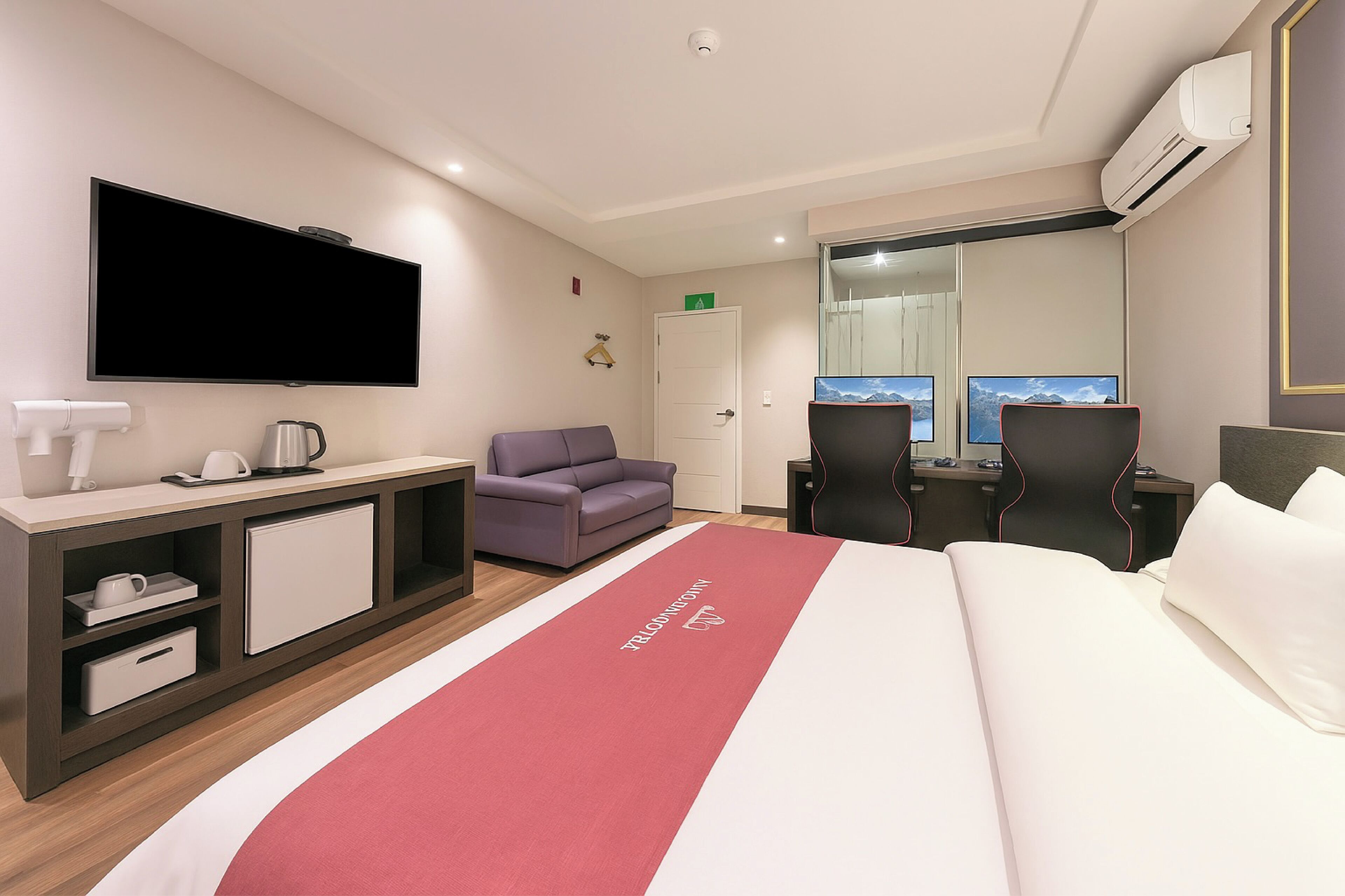 A hotel room with a large bed, a desk with a computer, a sofa, a TV mounted on the wall, and an air conditioning unit.