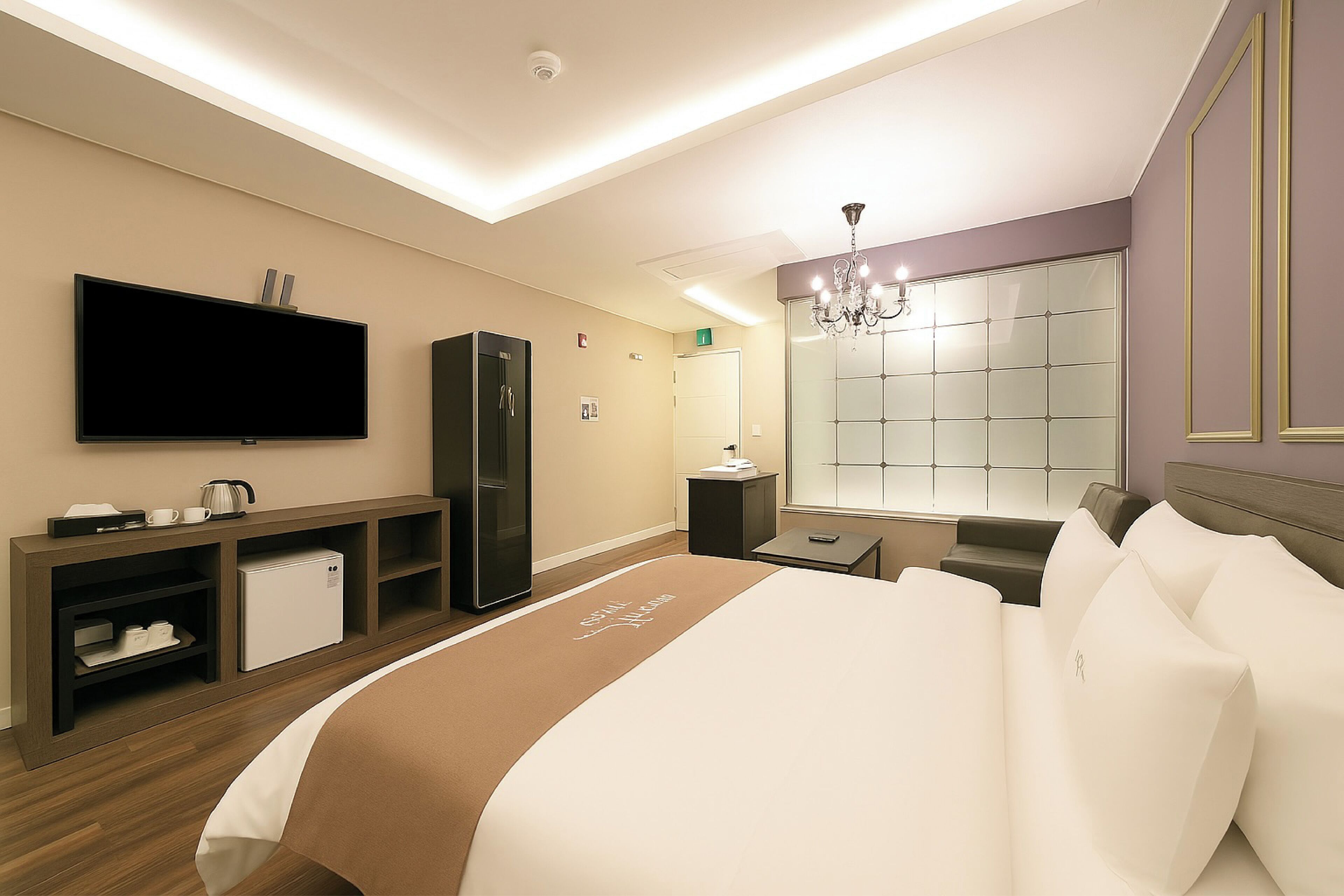 A modern hotel room with a large bed, a flat-screen TV, a minibar, a seating area, and a large window with a view.