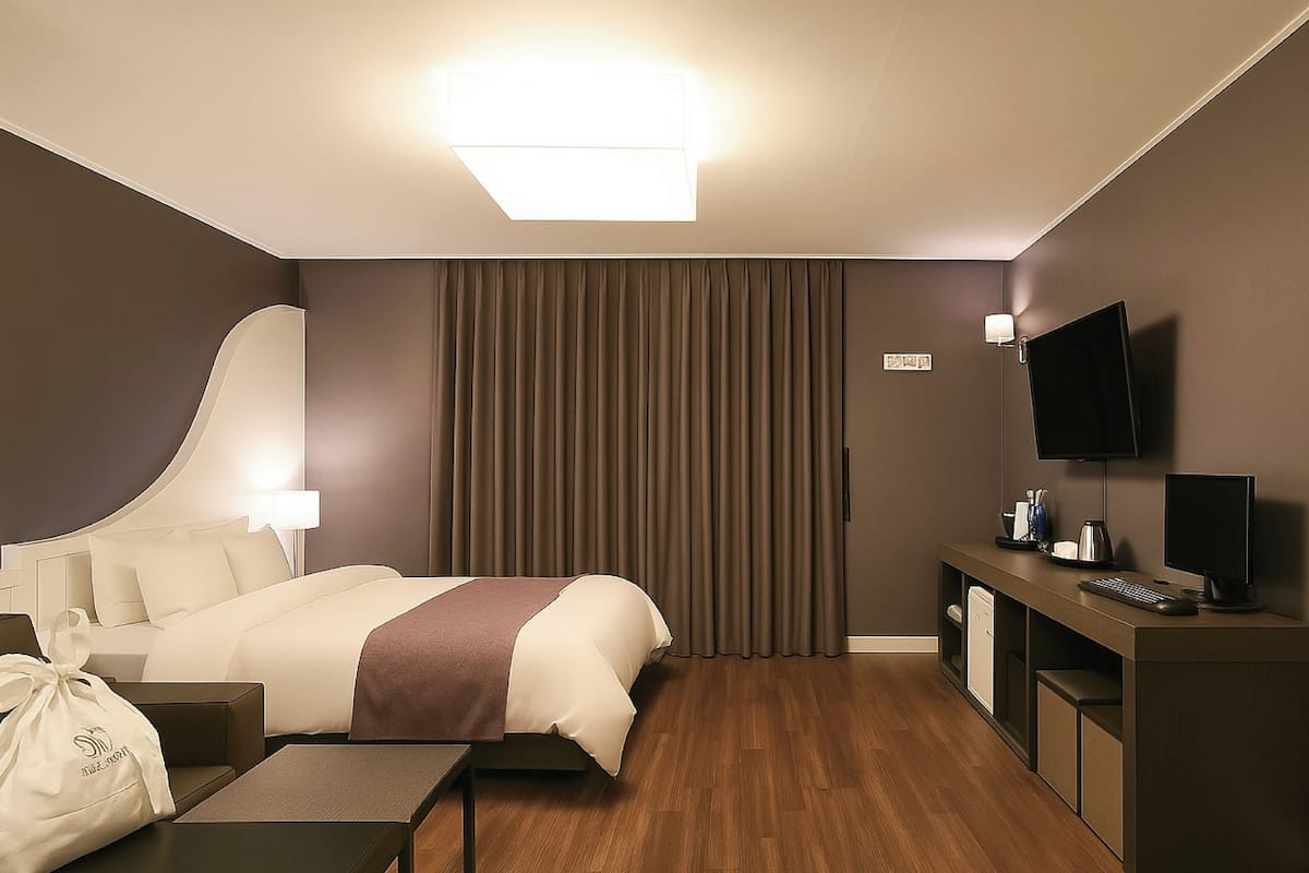 A hotel room with a bed, bedside table, lamp, TV, desk, and computer.