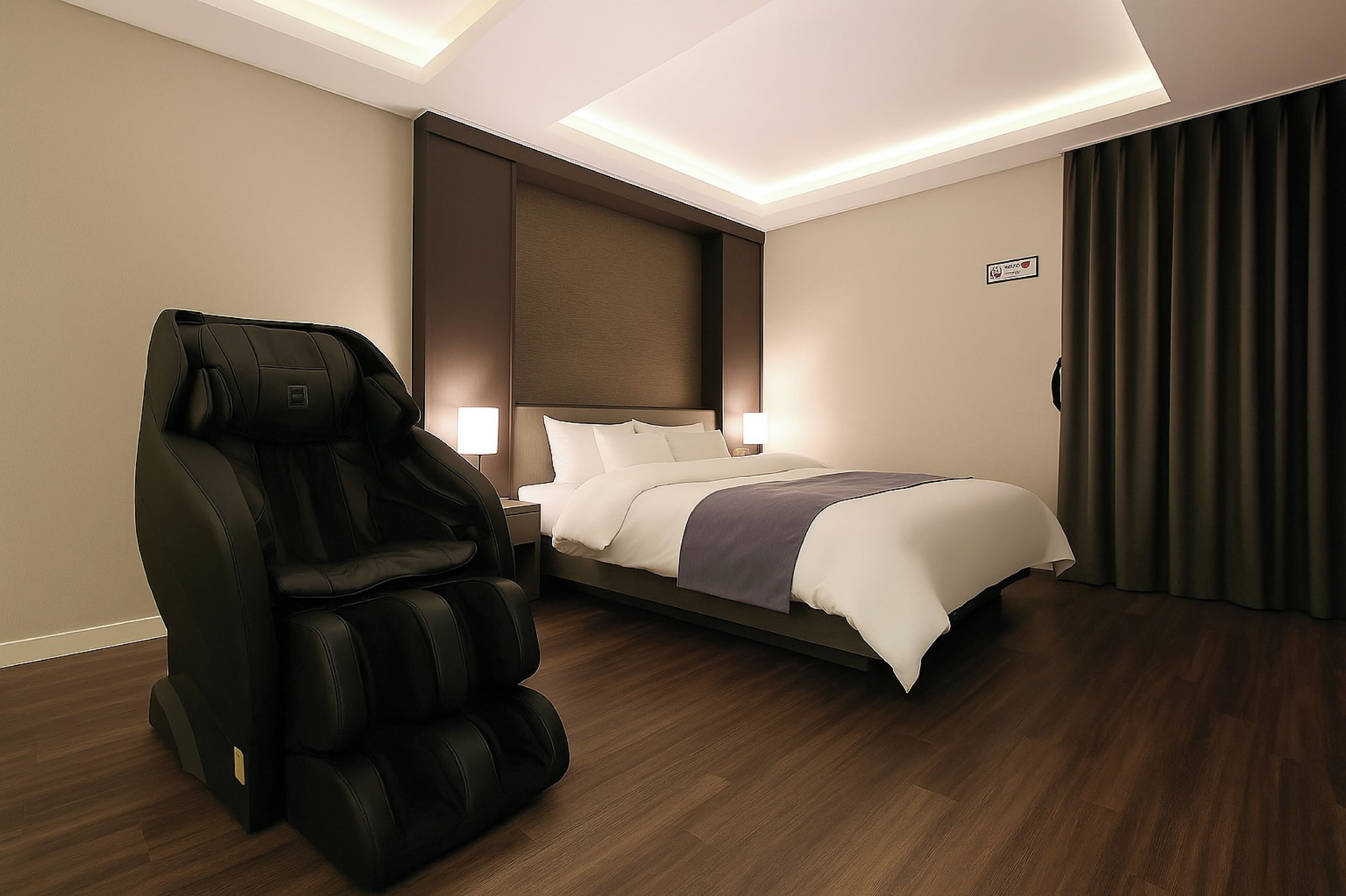 Ein Hotelzimmer mit Massagesessel, einem Bett mit weißer Bettwäsche, einem Nachttisch und einem an der Wand befestigten Schild.