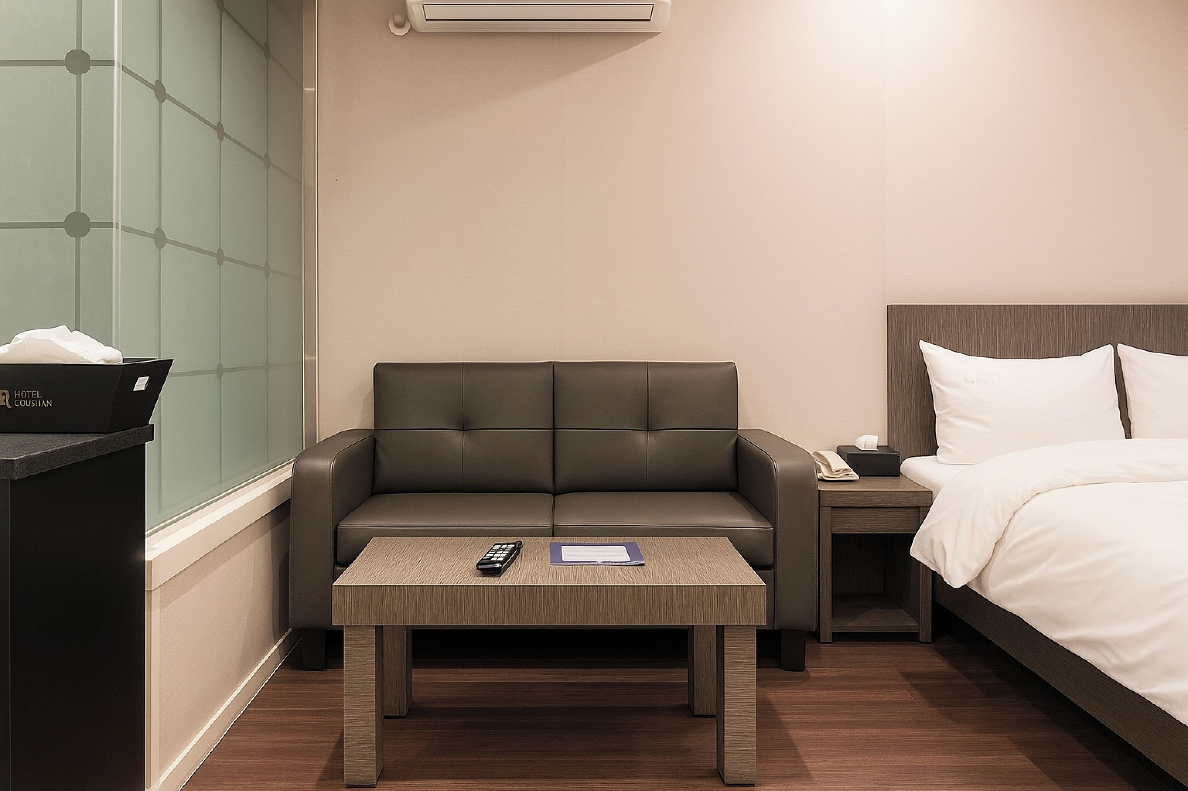 Habitación de hotel con sofá, una mesita con un control remoto y papeles, una mesita de noche con un teléfono y una caja, y una cama con ropa blanca.