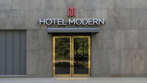 Moderni hotellin sisäänkäynti, jossa on lasiovi ja metallikehys. Oven yläpuolella on hotellin logo ja sen alla nimi ”HOTEL MODERN”.