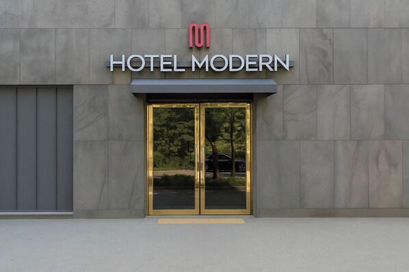 Una entrada de hotel moderna con puerta de vidrio y estructura metálica, que muestra un logotipo sobre el nombre "HOTEL MODERN".