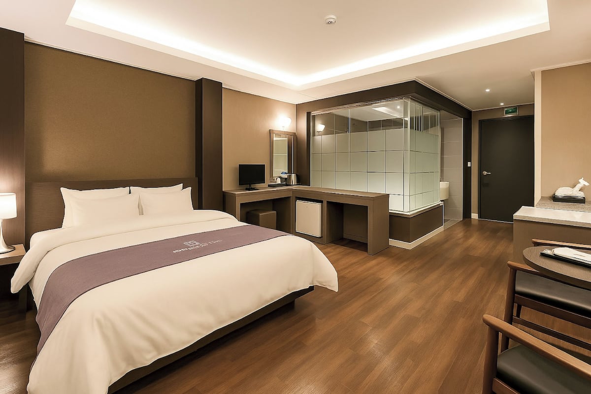 A modern hotel room with a large bed, a desk, a glass shower enclosure, and a small seating area.