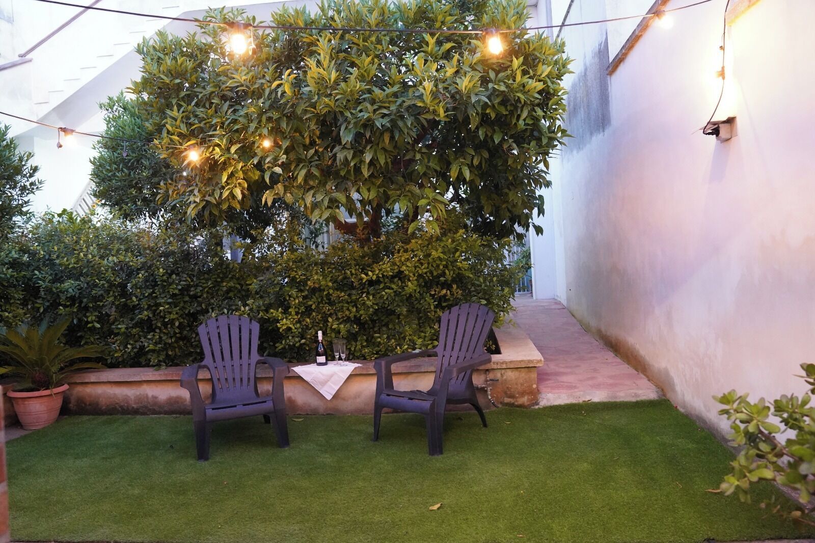 An outdoor seating area with two chairs and a table, surrounded by greenery and string lights.