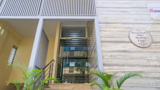 Una entrada moderna a un edificio con puerta de vidrio, un letrero que indica 'Purnachandra Billva Residency #1667', y una pared de madera con una placa.