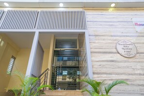 A modern building entrance with a glass door, a sign indicating 'Purnachandra Billva Residency #1667', and a wooden wall with a plaque. - Housr Charlie 1 BHK Apartment Bangalore (Bengaluru)