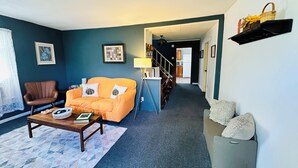 A living room with a sofa, armchair, coffee table, and a staircase leading to a kitchen. - Pet friendly Near Lake George and Saratoga (Corinth)