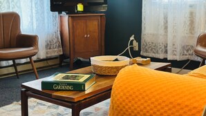 A room with a television on a wooden cabinet, two chairs, a table with a book, and a basket. - Pet friendly Near Lake George and Saratoga (Corinth)
