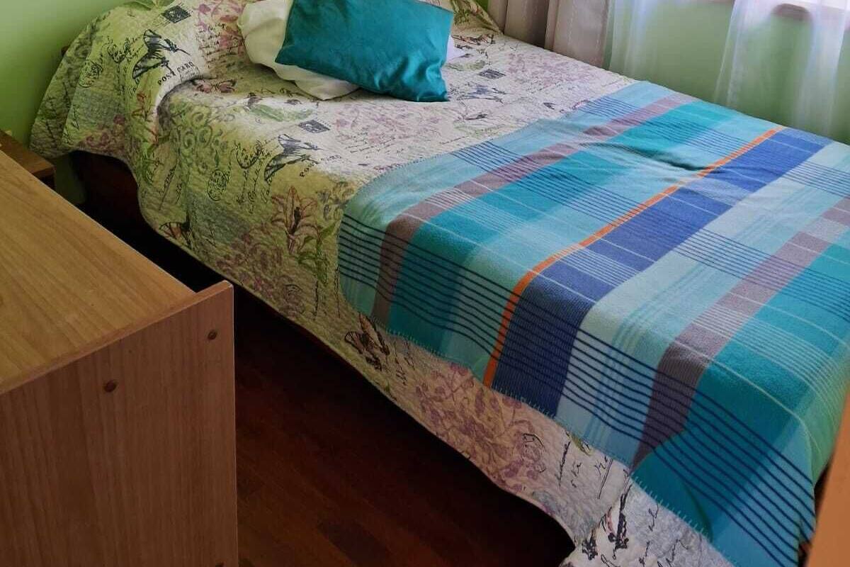 A single bed with a floral bedspread and a blue pillow, a wooden nightstand, and a window with sheer curtains.