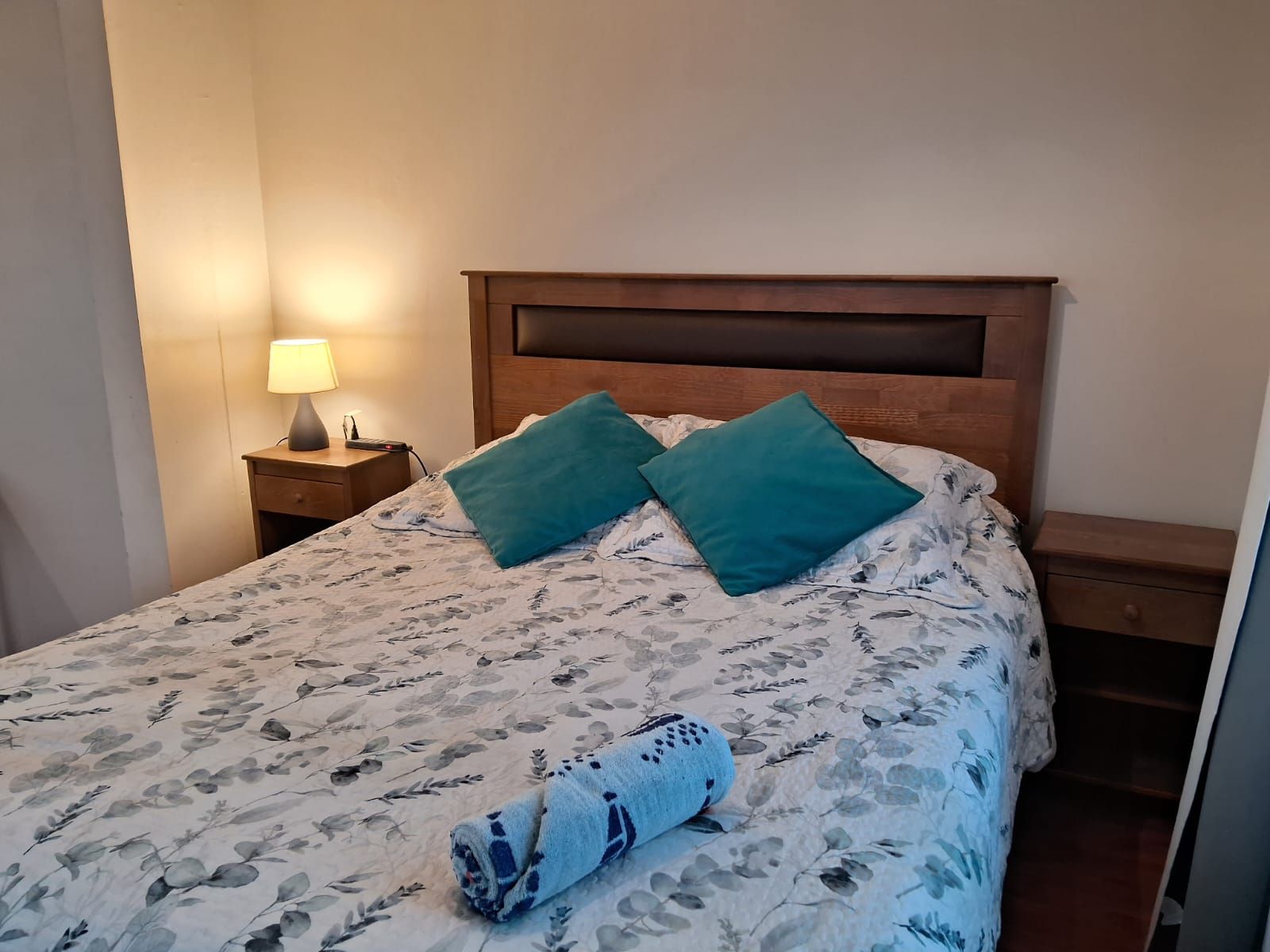 Una cama bien hecha con ropa de cama floral, dos almohadas color turquesa y una toalla enrollada.