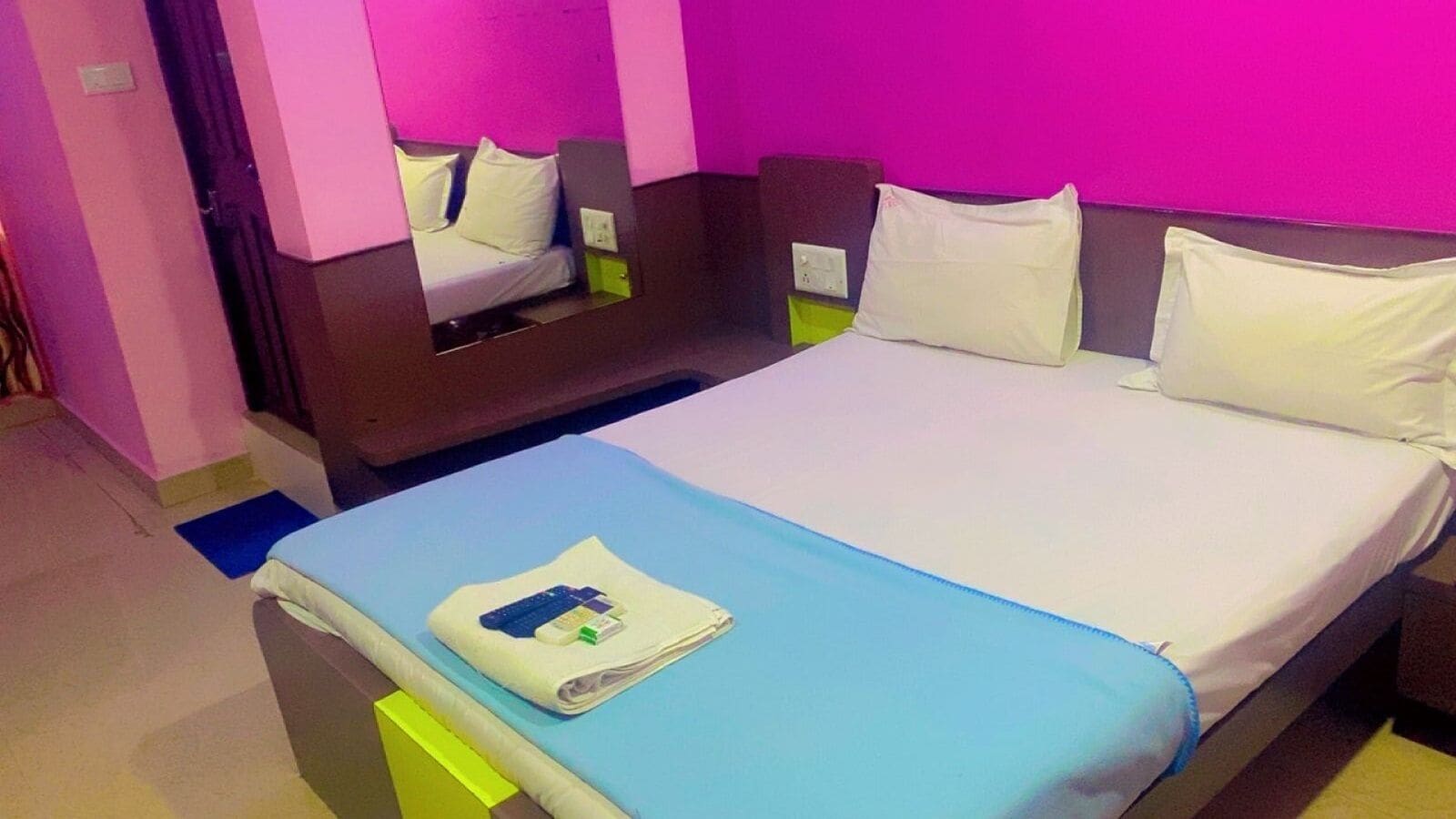 Et hotelværelse med en seng, et natbord, et spejl og en væg i neonrosa.