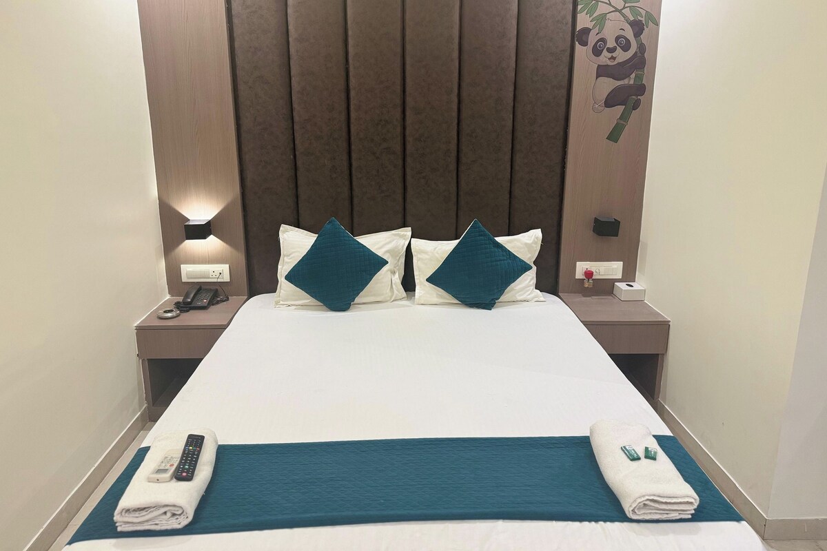 A hotel room with a large bed, two pillows, a bedside table with a lamp, a phone, and a remote control on the bed.