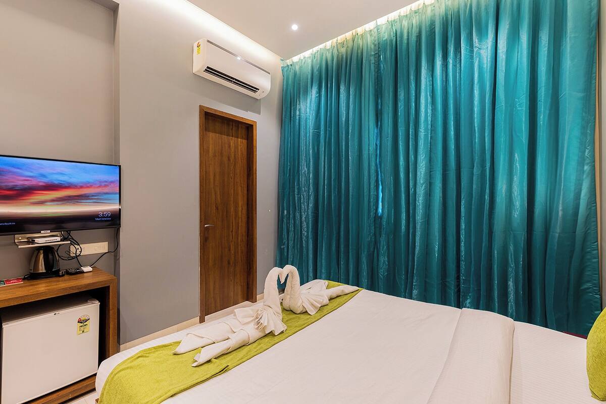 A hotel room with a bed, a television on a wooden stand, a small refrigerator, and teal curtains.