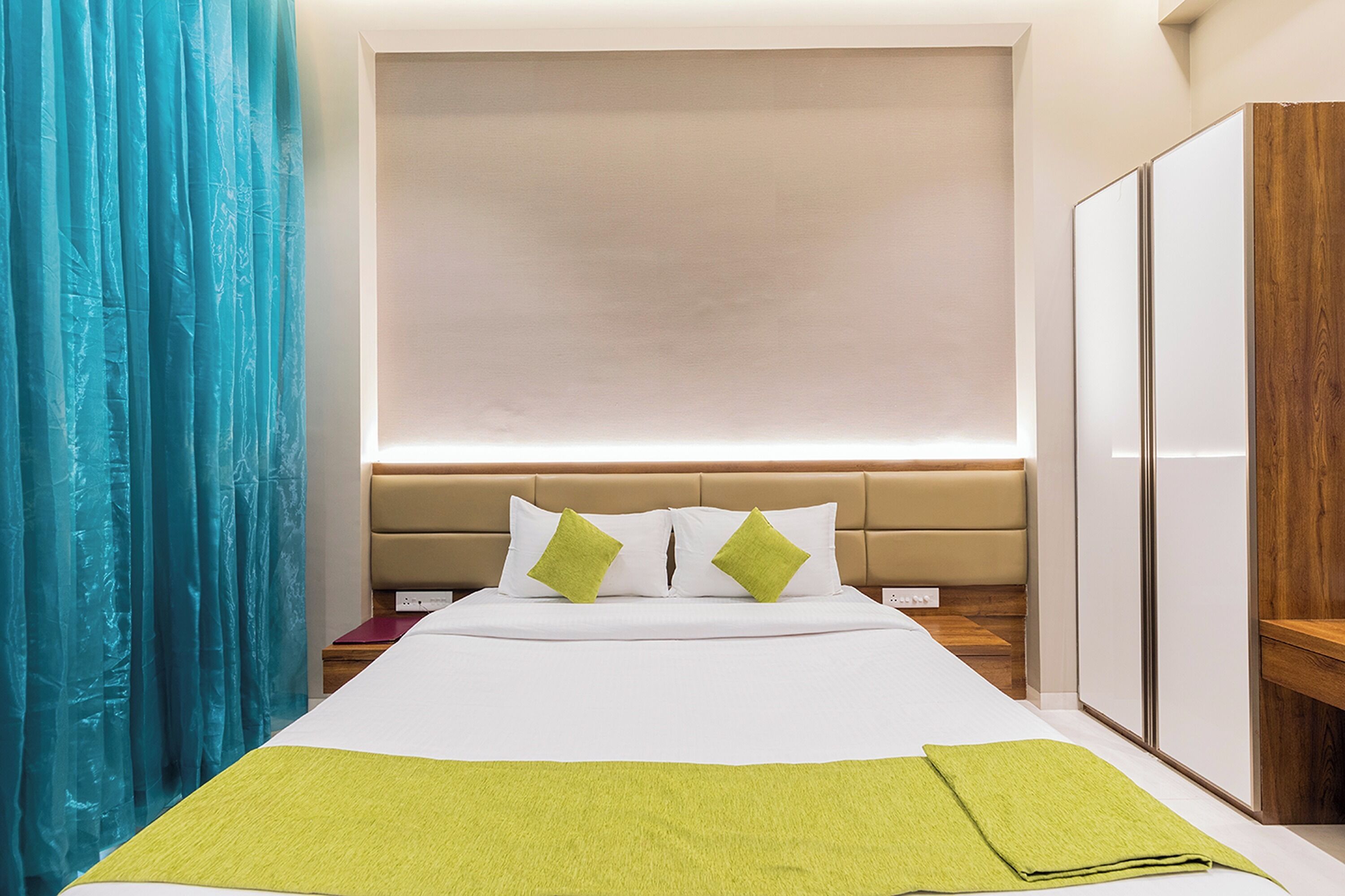 Une chambre d’hôtel avec un grand lit, une tête de lit agrémentée de deux coussins verts, une armoire en bois et un rideau bleu.