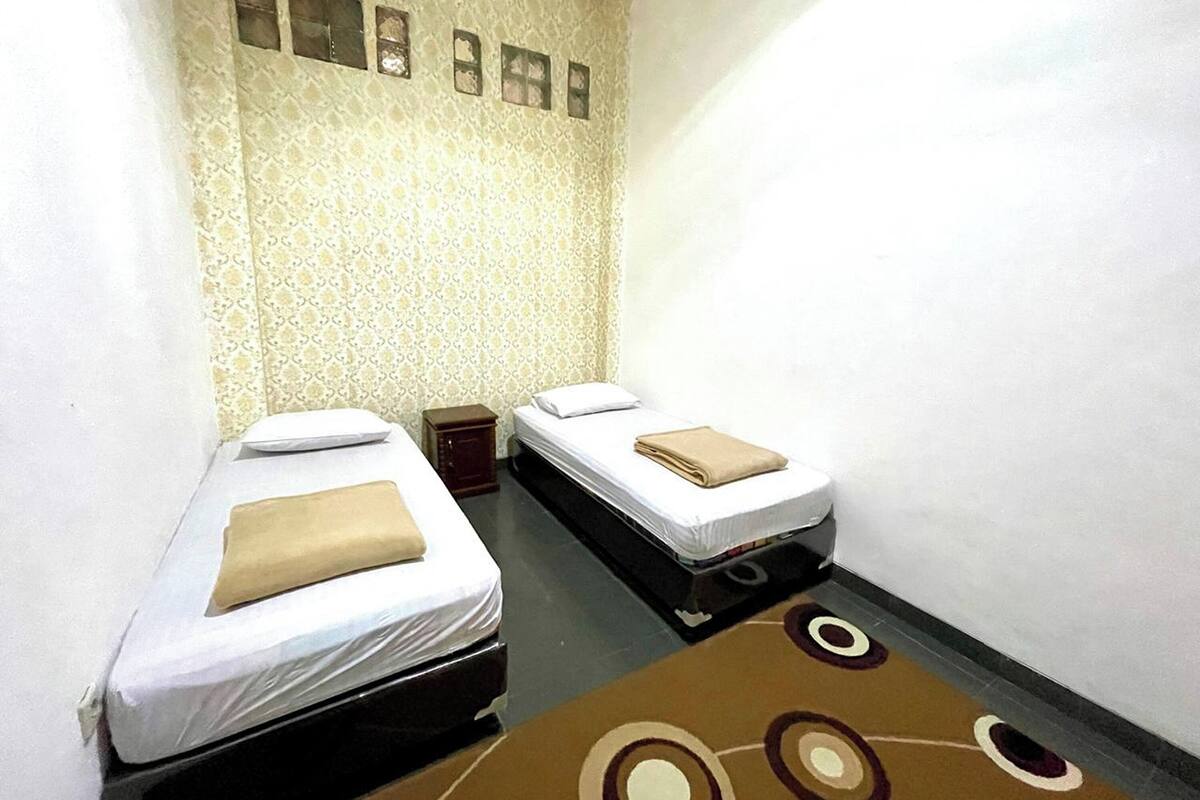 A small, narrow room with two beds, a patterned wallpaper, and a brown carpet with circular designs.