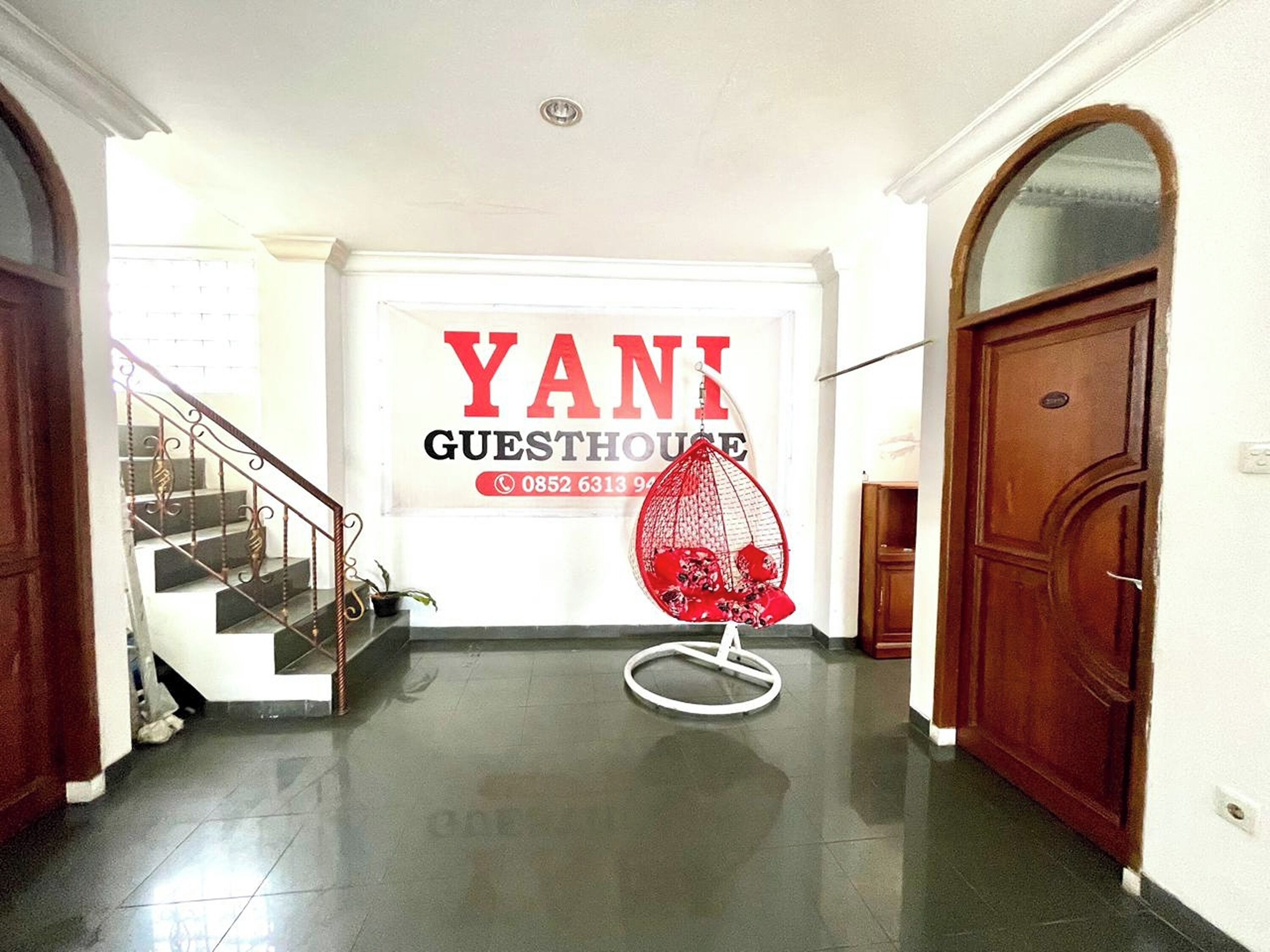 A hallway with a staircase, a red hanging chair, and a sign for 'YANI GUESTHOUSE' with a phone number.