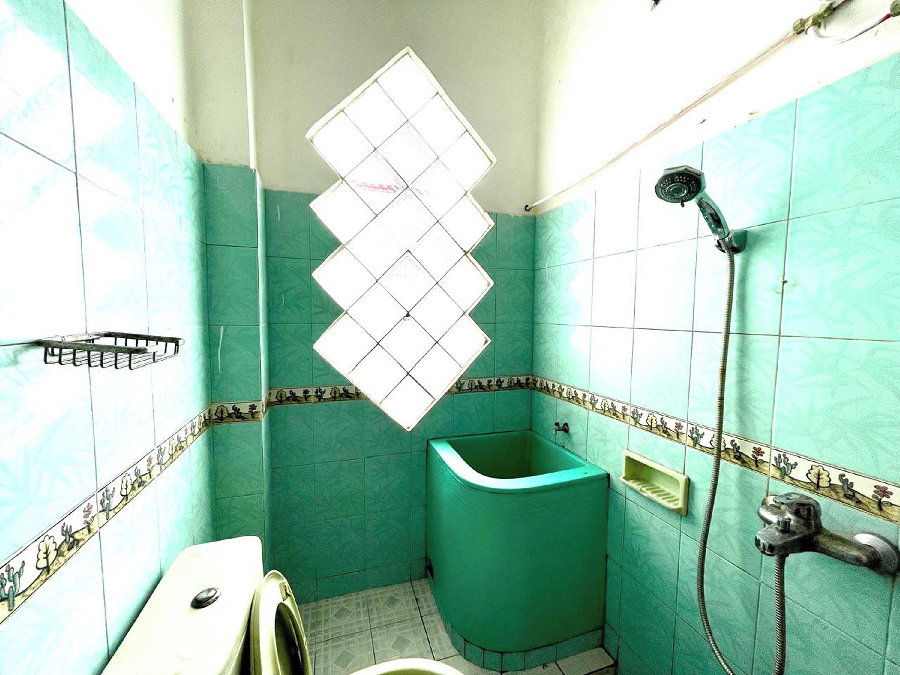 Ein Badezimmer mit türkisfarbenen Fliesen, einer Dusche, einer Toilette und einem Fenster mit Rautenmuster.