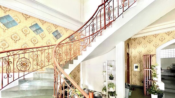 A grand staircase with ornate metal railings, leading to a landing with a white shelf holding potted plants. - Yani Guesthouse (Bukittinggi)