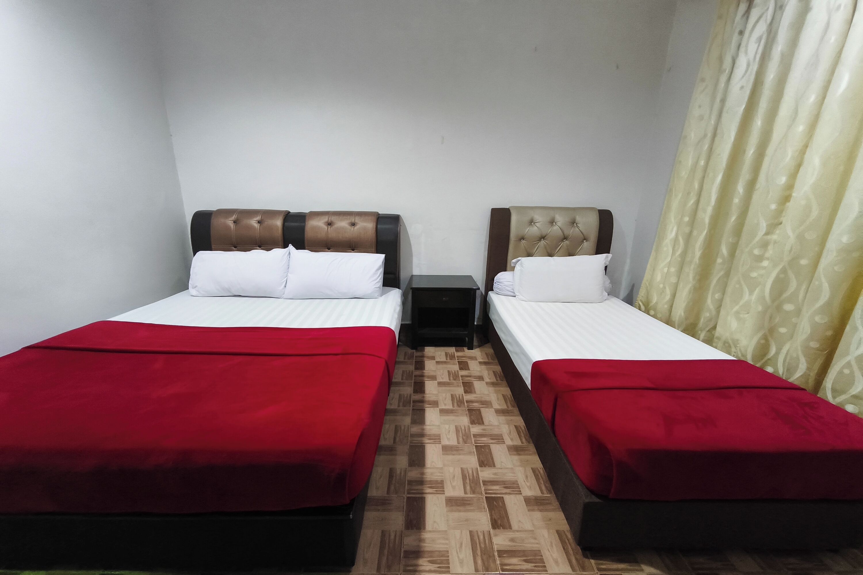 Ein Zimmer mit zwei Betten, jeweils mit roter Bettdecke und weißen Kissen. Die Betten haben gesteppte Kopfteile in Braun- und Beigetönen. Zwischen den Betten befindet sich ein kleiner Nachttisch. Der Boden ist Holzdielenboden mit Fischgrätmuster.