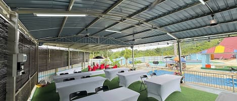An outdoor covered area with tables and chairs, artificial grass, and a view of a playground and buildings in the background.