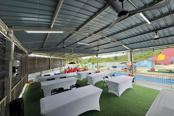 An outdoor covered area with tables and chairs, artificial grass, and a view of a playground and buildings in the background.
