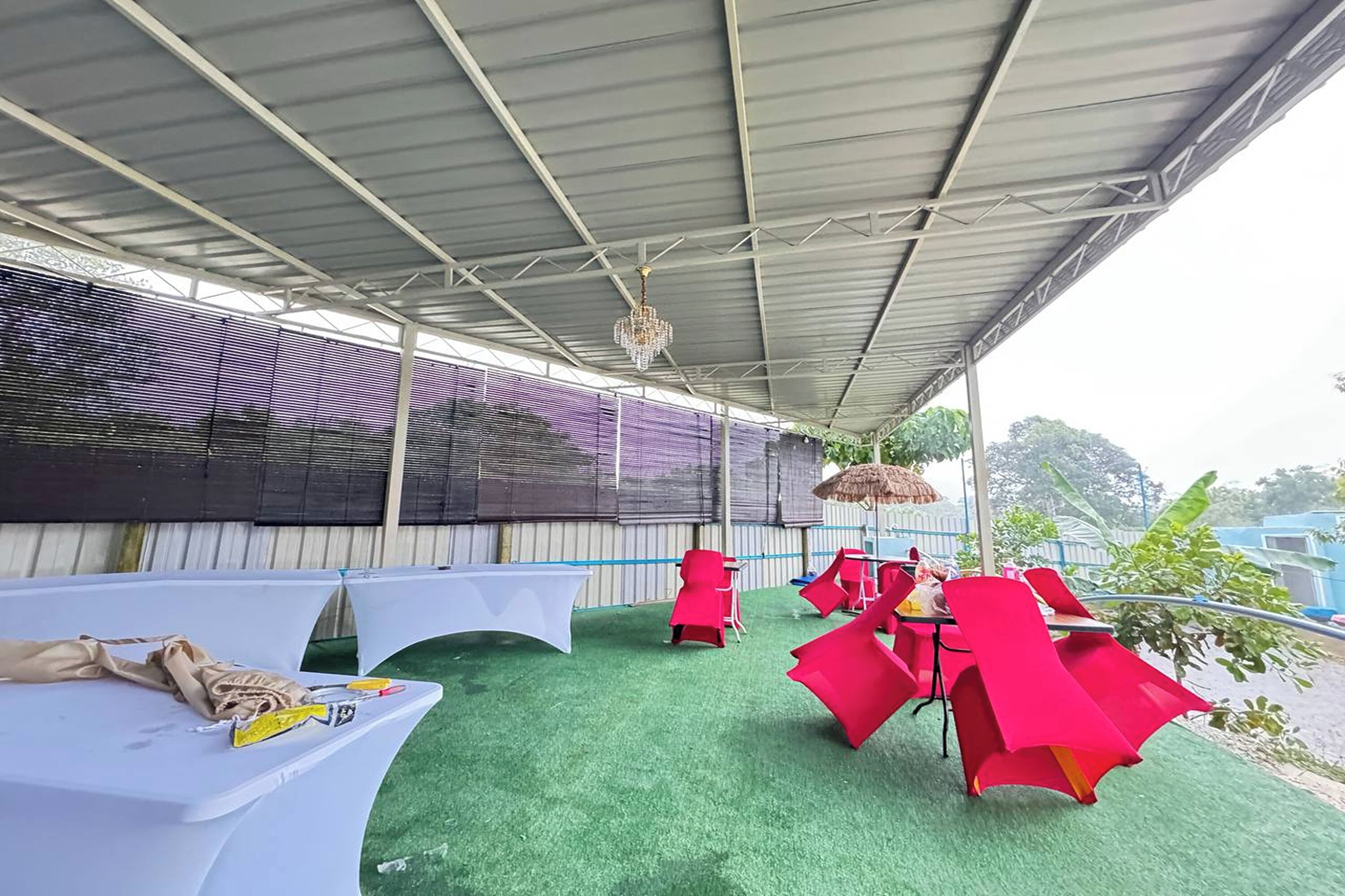 An outdoor covered area with tables and chairs, a hanging chandelier, and a view of trees and buildings in the background.